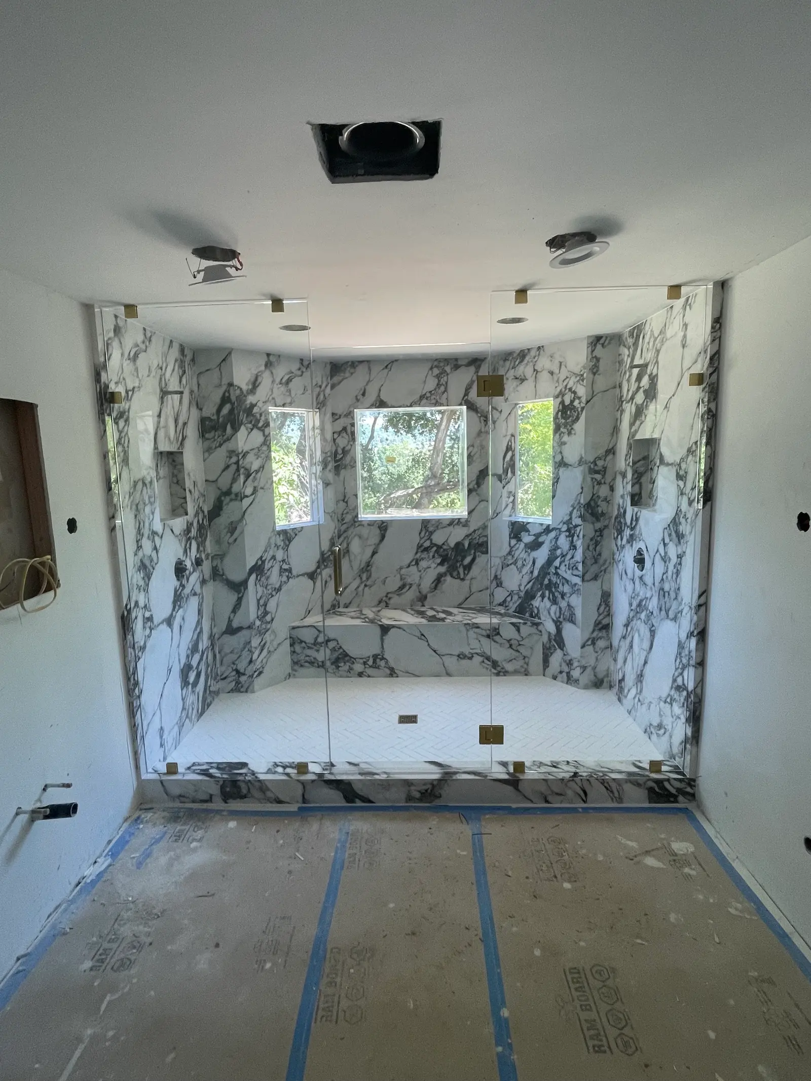 Wide walk-in shower with frameless glass panels, brass clips and bookmatched marble around three windows