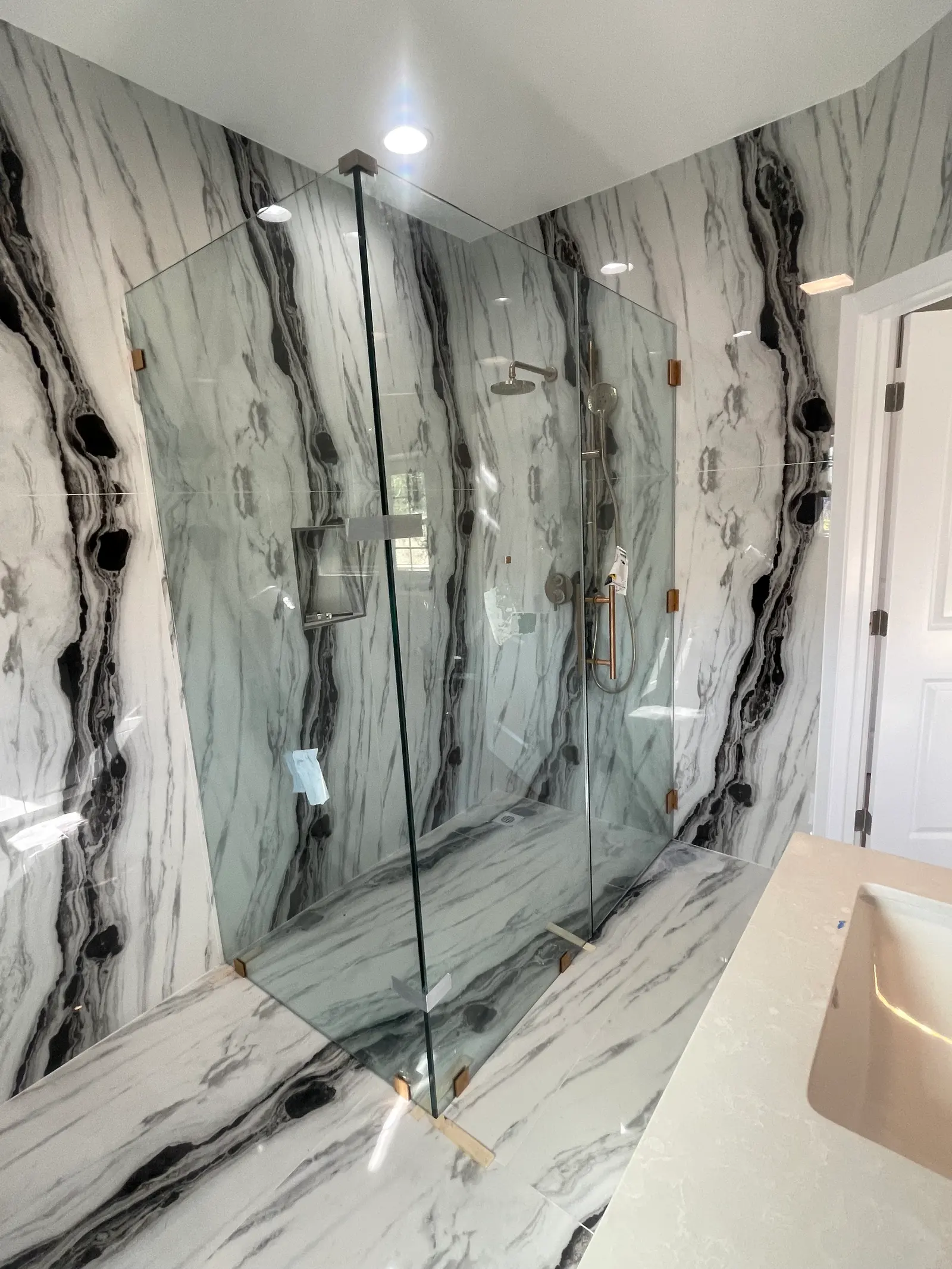 Frameless corner shower enclosure with brushed-brass clips against bookmatched black-and-white quartzite