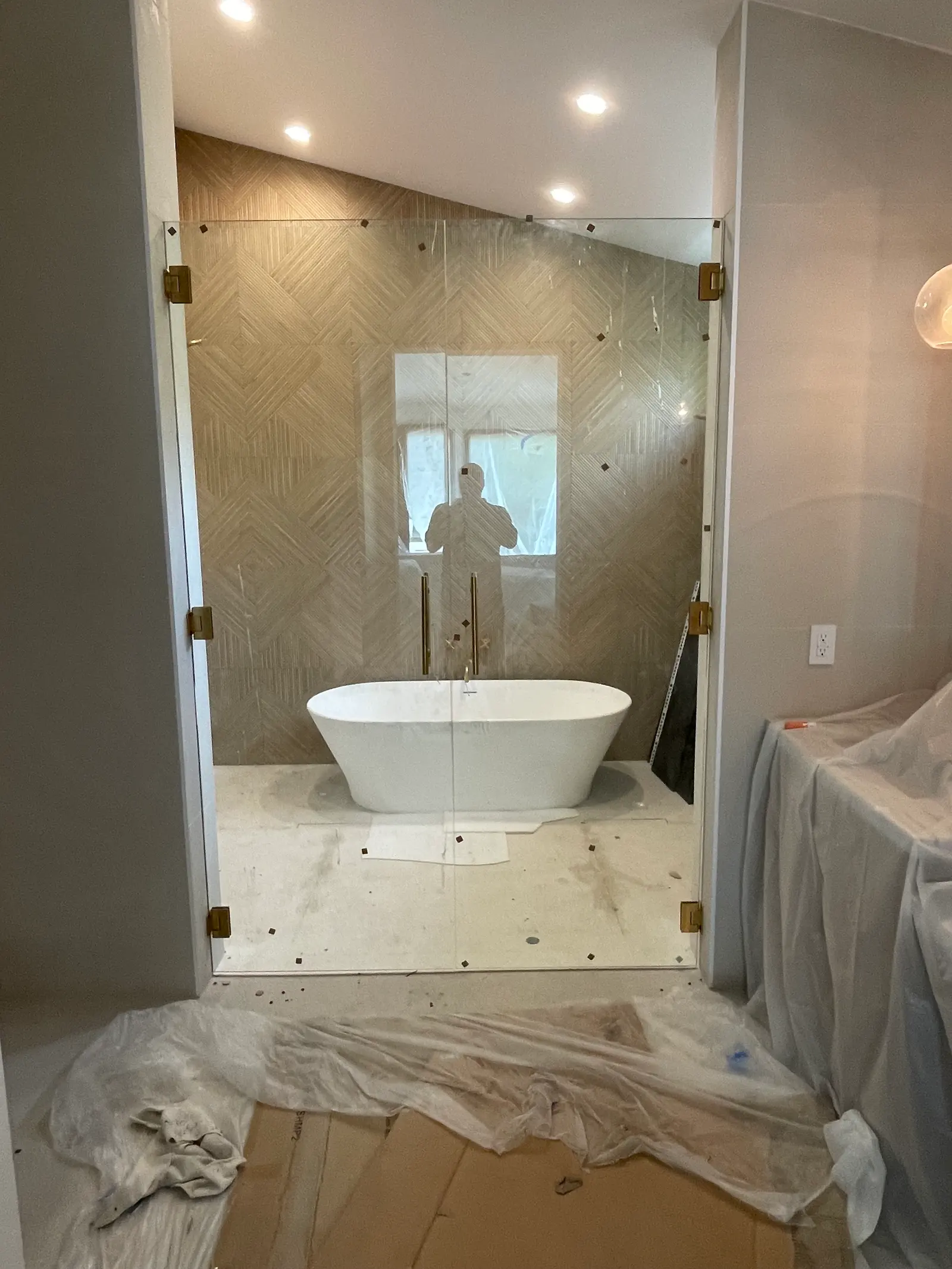 Floor-to-ceiling glass panels with brass hinges enclosing a freestanding tub against herringbone tile