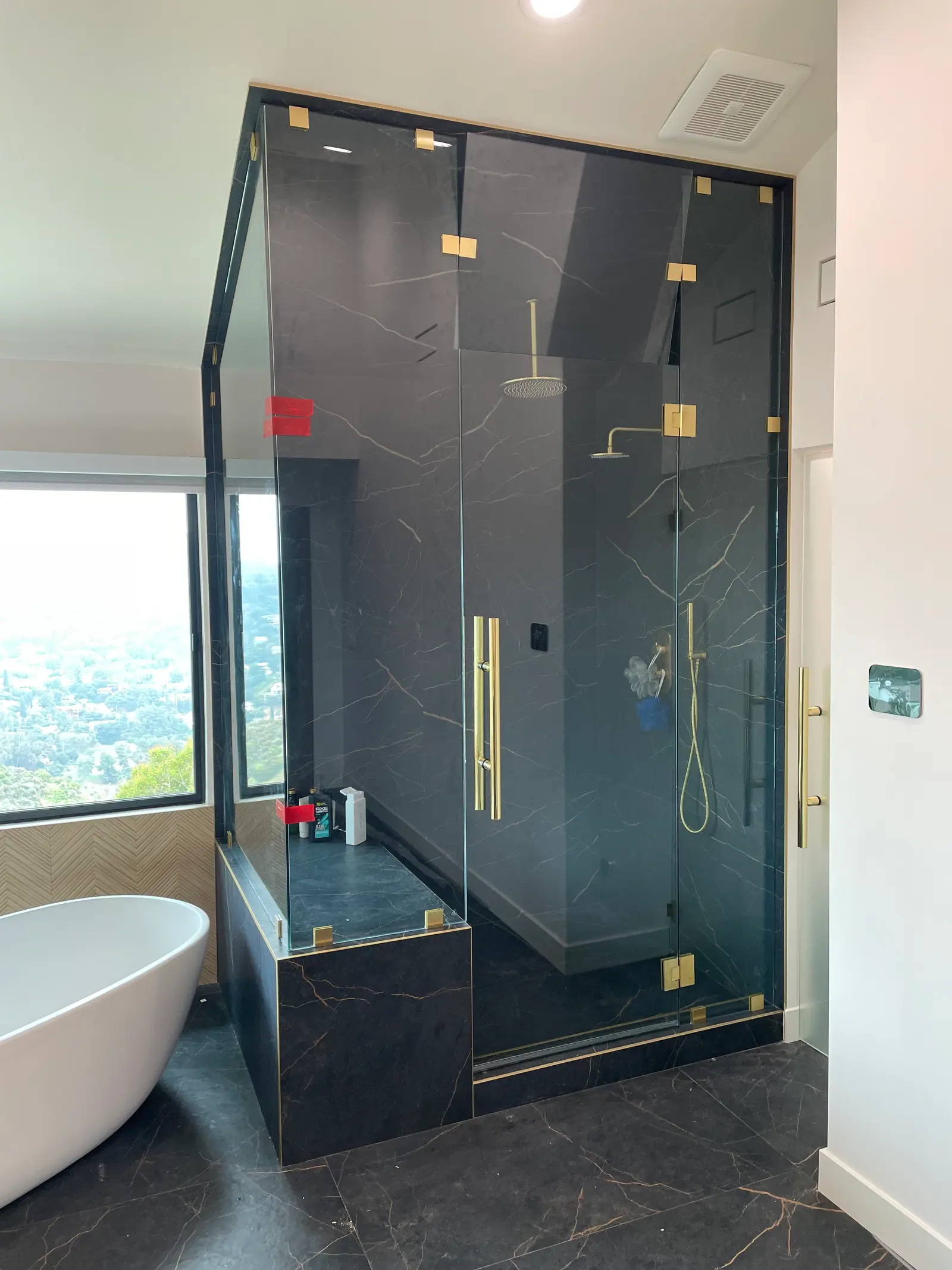 Black marble steam shower with brushed-brass hardware beside a freestanding tub and a hillside window
