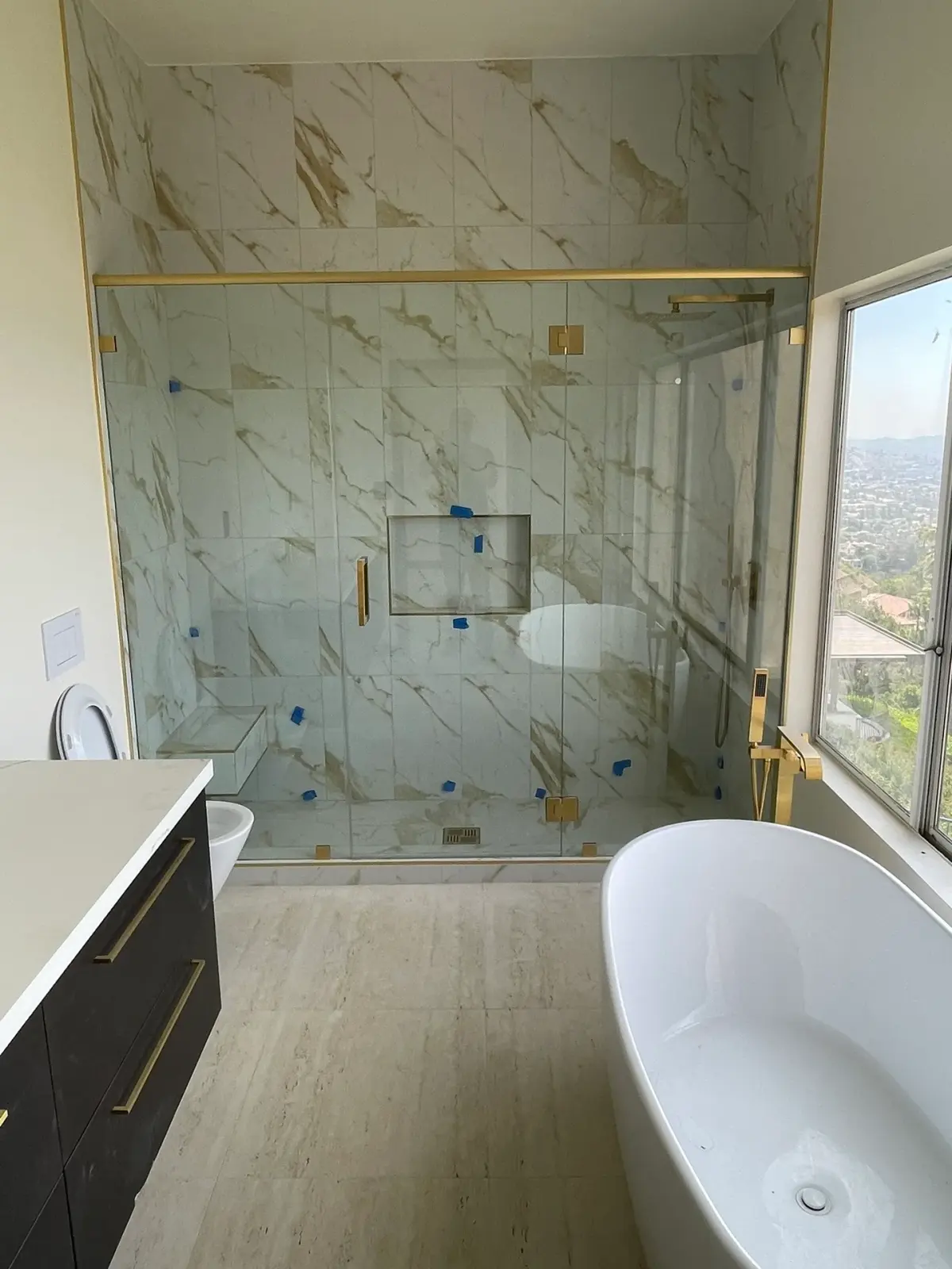 Gold-framed glass shower enclosure beside a freestanding tub with hillside views of Los Angeles